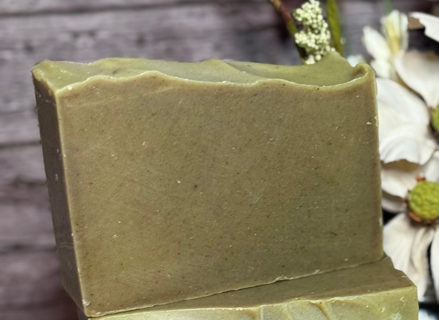 Green bar of soap on a wooden surface with blurred flowers in the background