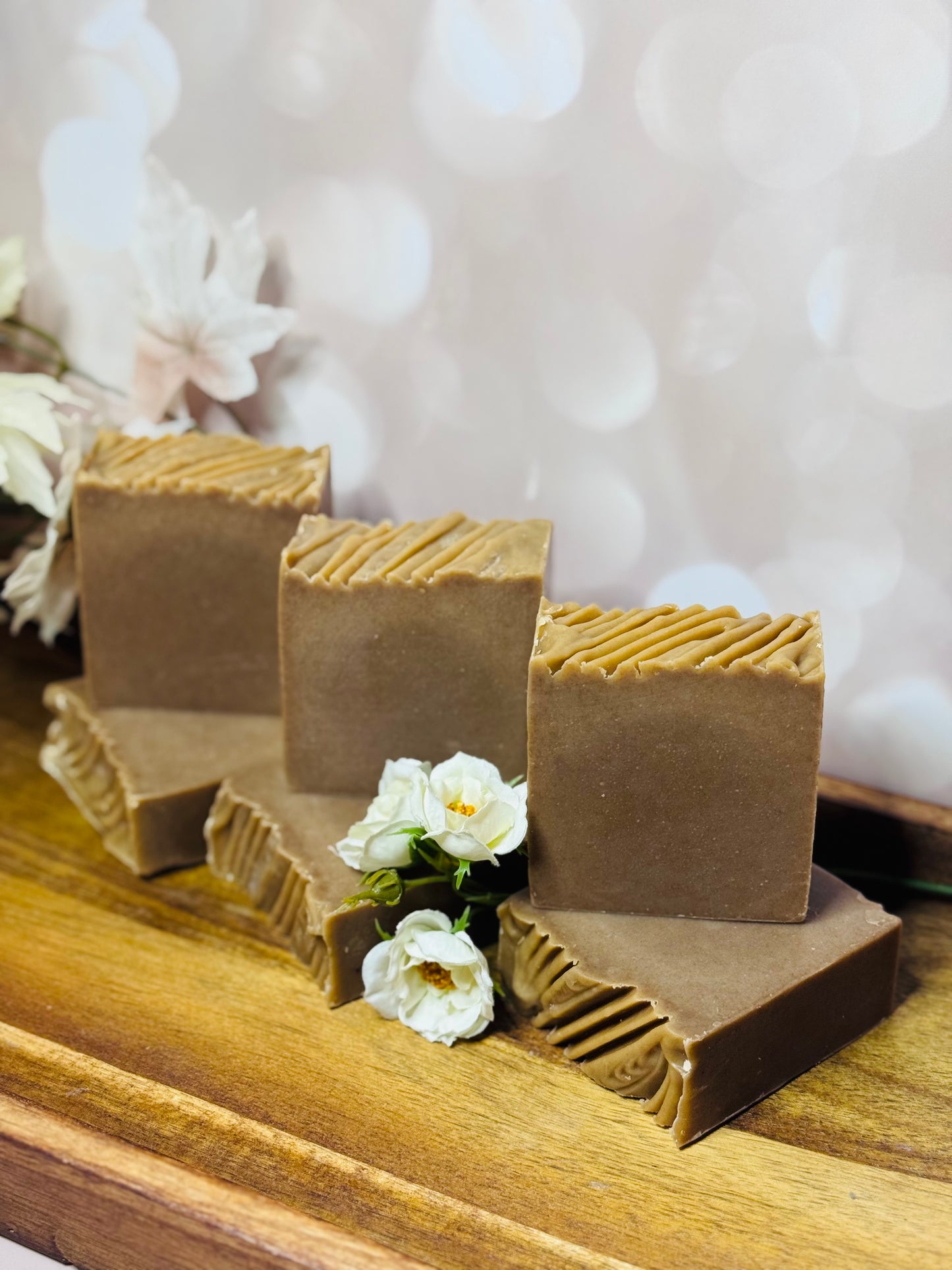 Six square brown soap bars on a wooden surface with white flowers in the background