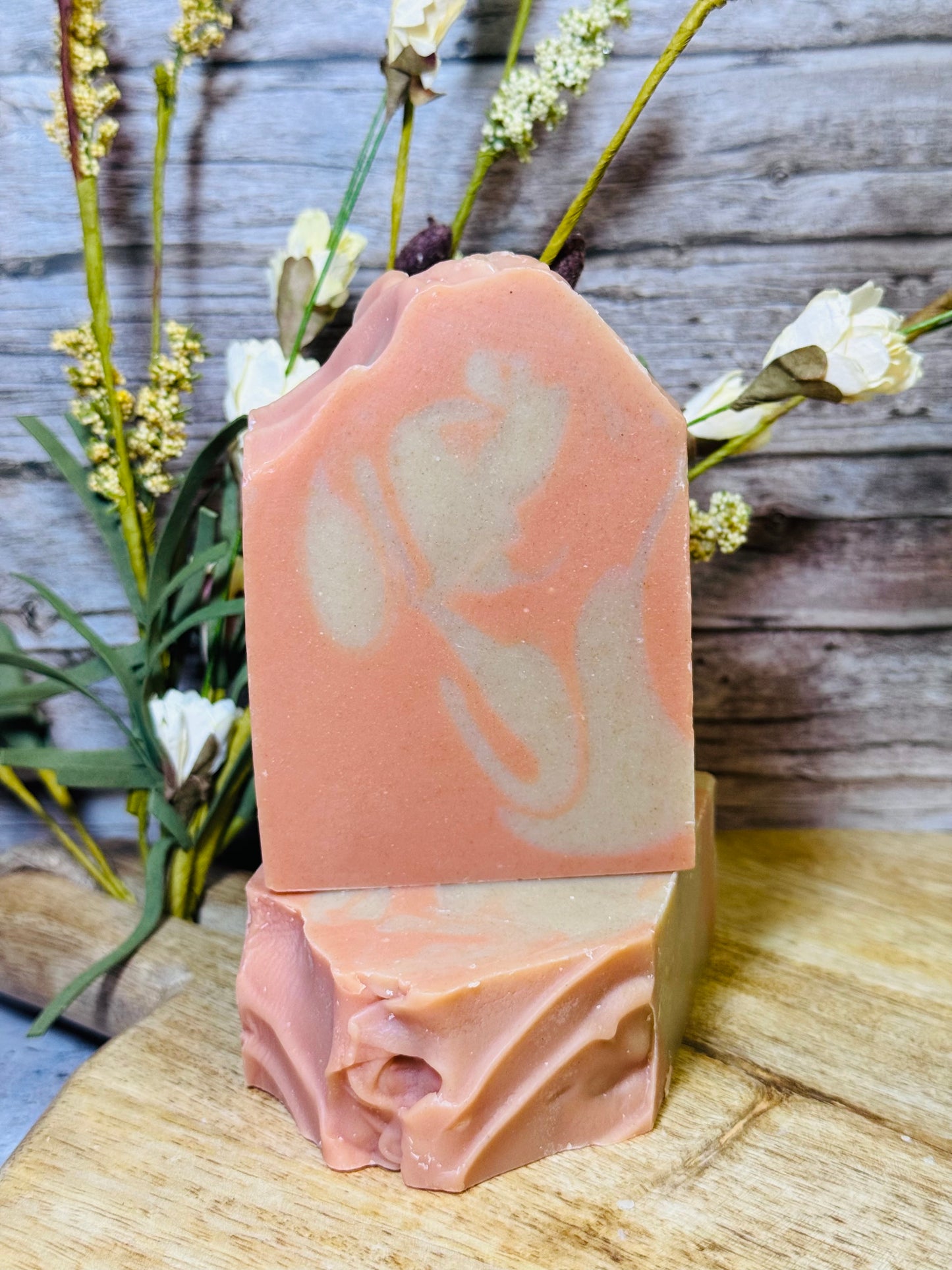 a Bar of soap a top of another bar of soap with a swirl design on a wooden surface with flowers in the background