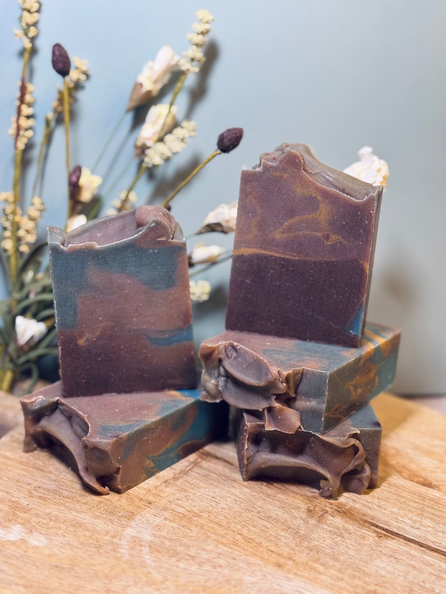 Handmade soap bars with natural texture on a wooden surface with dried plants in the background.