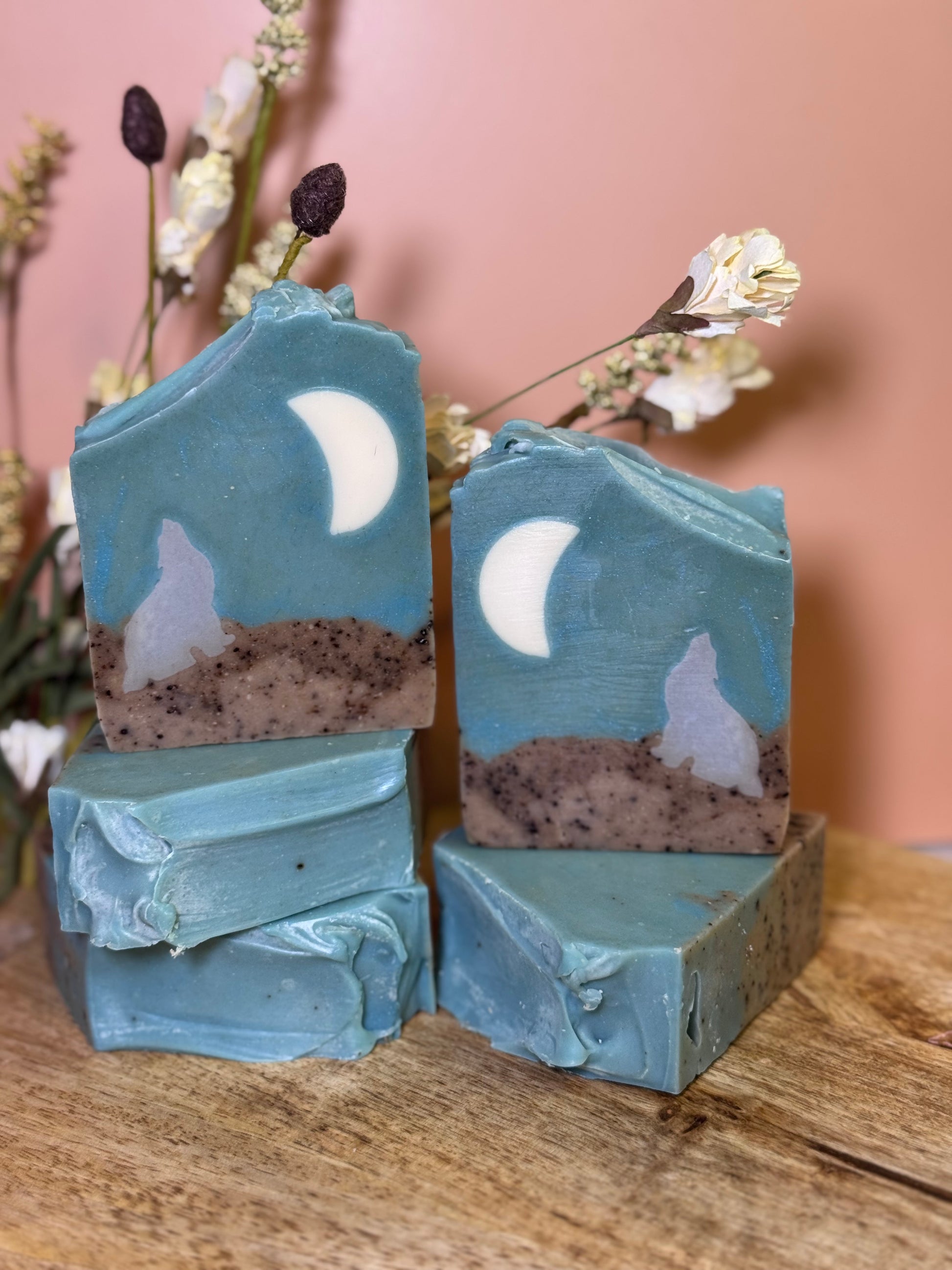 Five soap bars with moon and wolf designs on a wooden surface with flowers in the background.