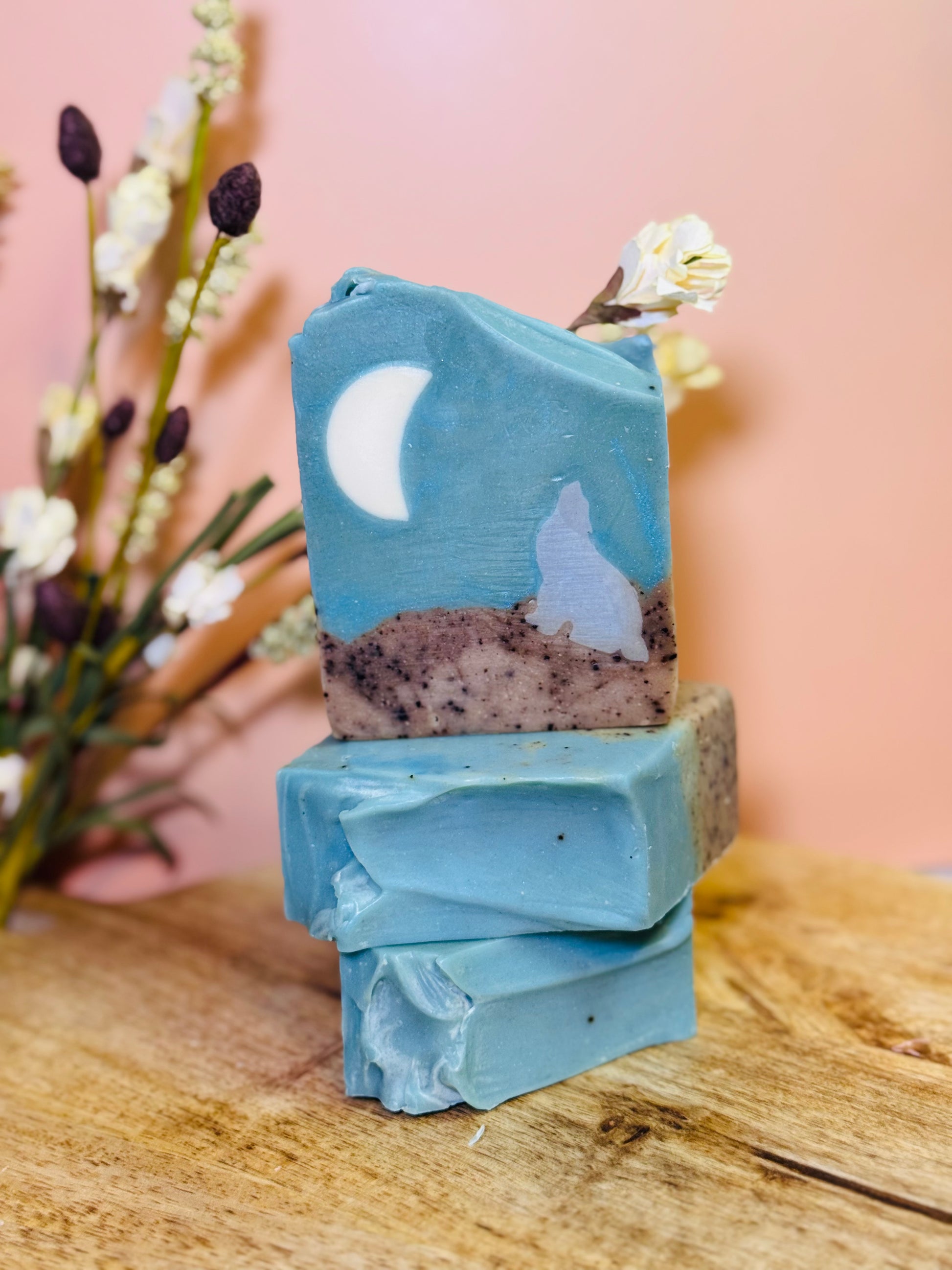 Stack of blue soap bars with a moon and wolf design on a wooden surface with flowers in the background.