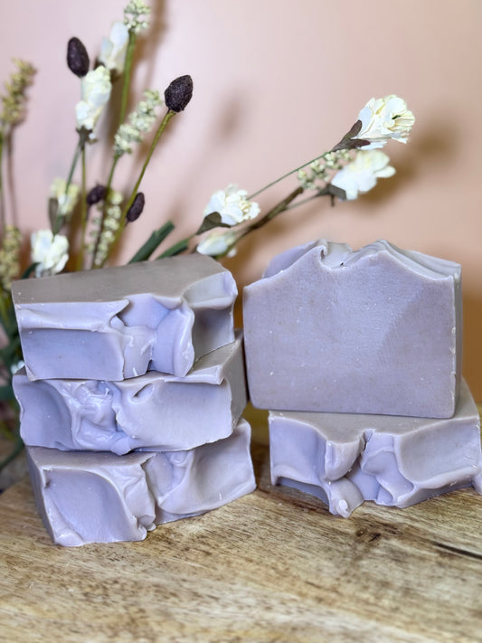 Stack of lavender-colored soap bars on a wooden surface with flowers in the background