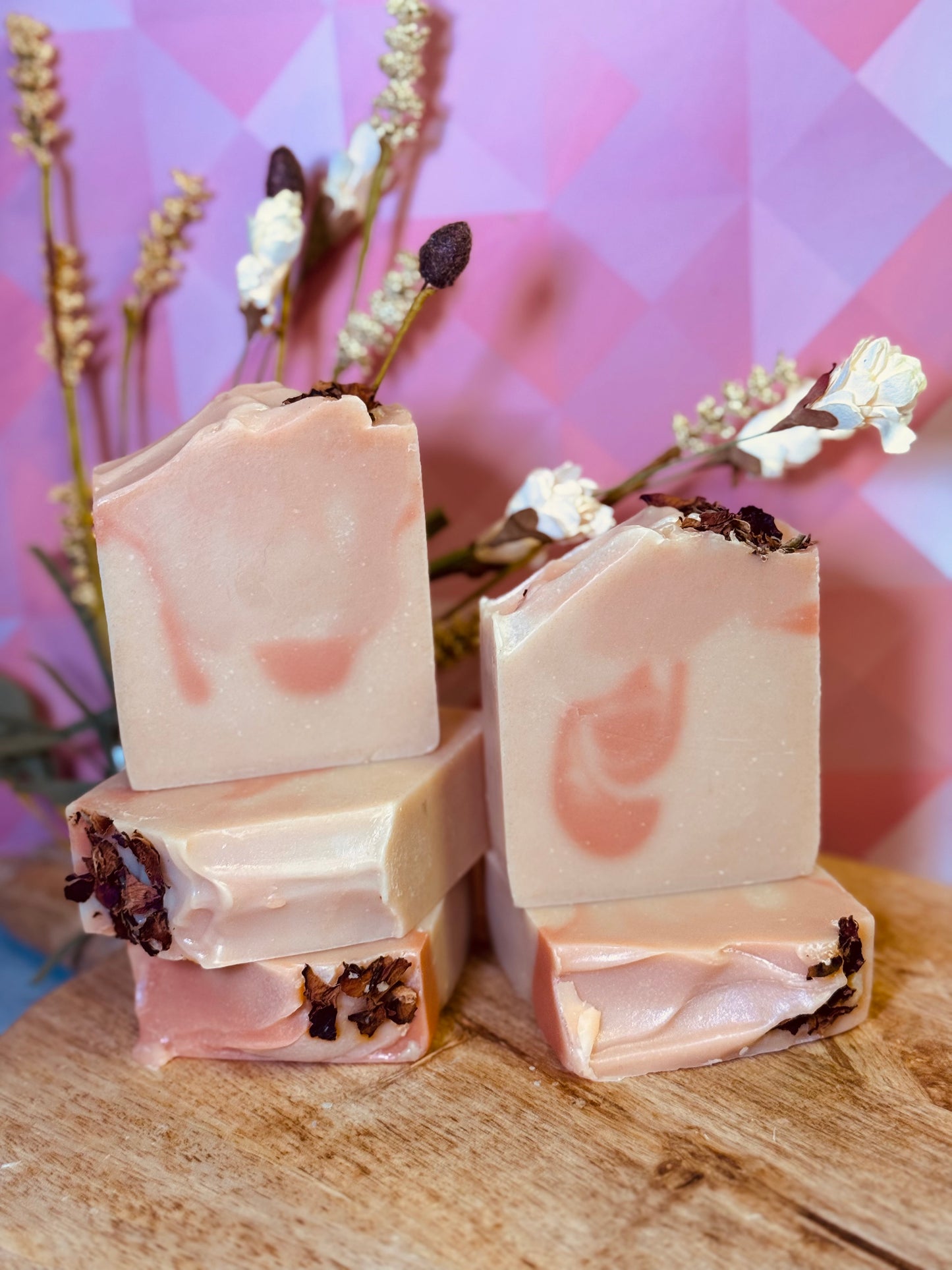 five bars of soap with floral elements on a wooden surface against a pink geometric background